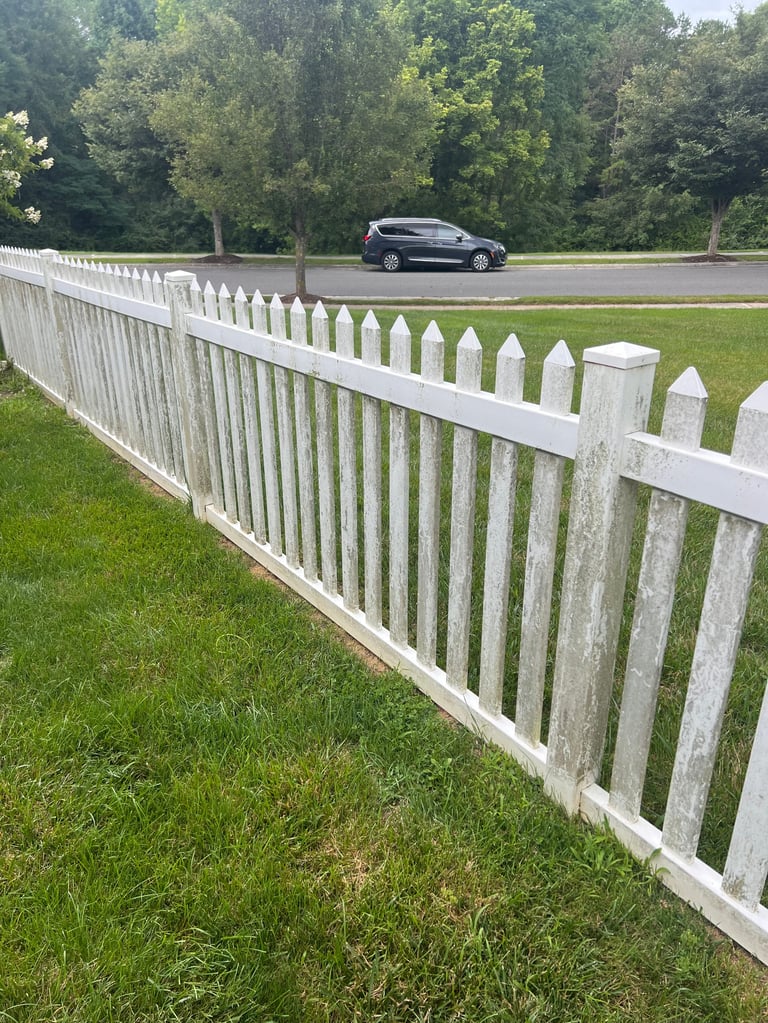 White picket fence along green lawn with car on asphalt road and tree-lined background
