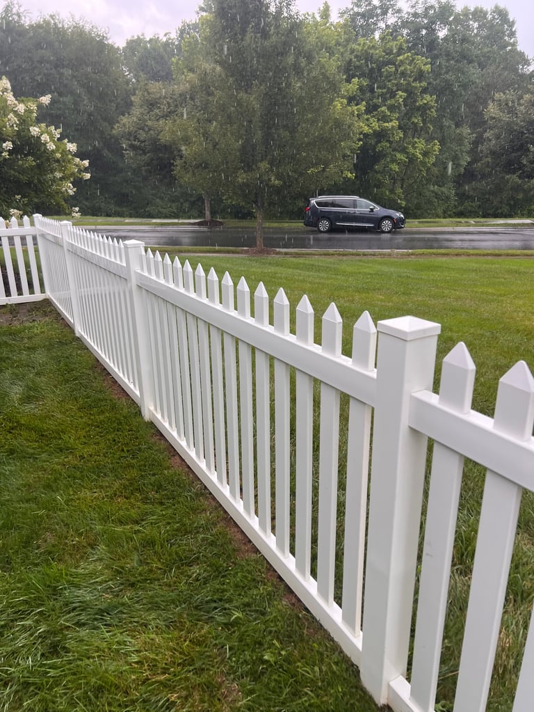 White picket fence along green lawn with dark car parked on road and trees in background