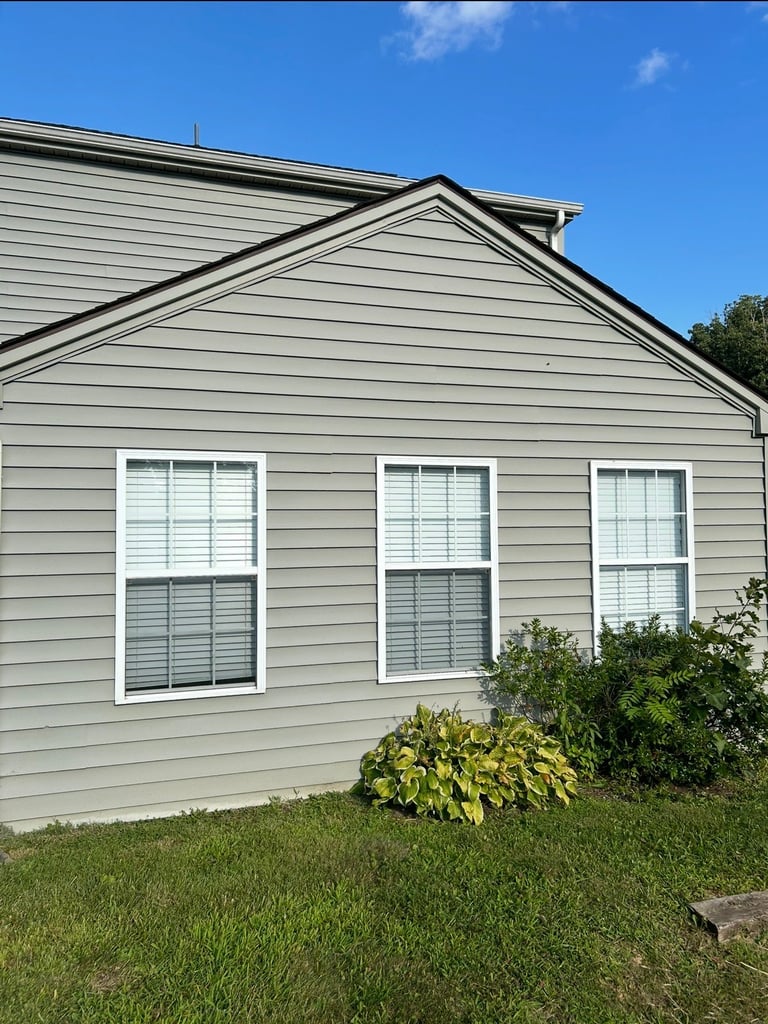 Front view of a gray vinyl-sided house with three windows, green lawn, and shrubs under a clear blue sky