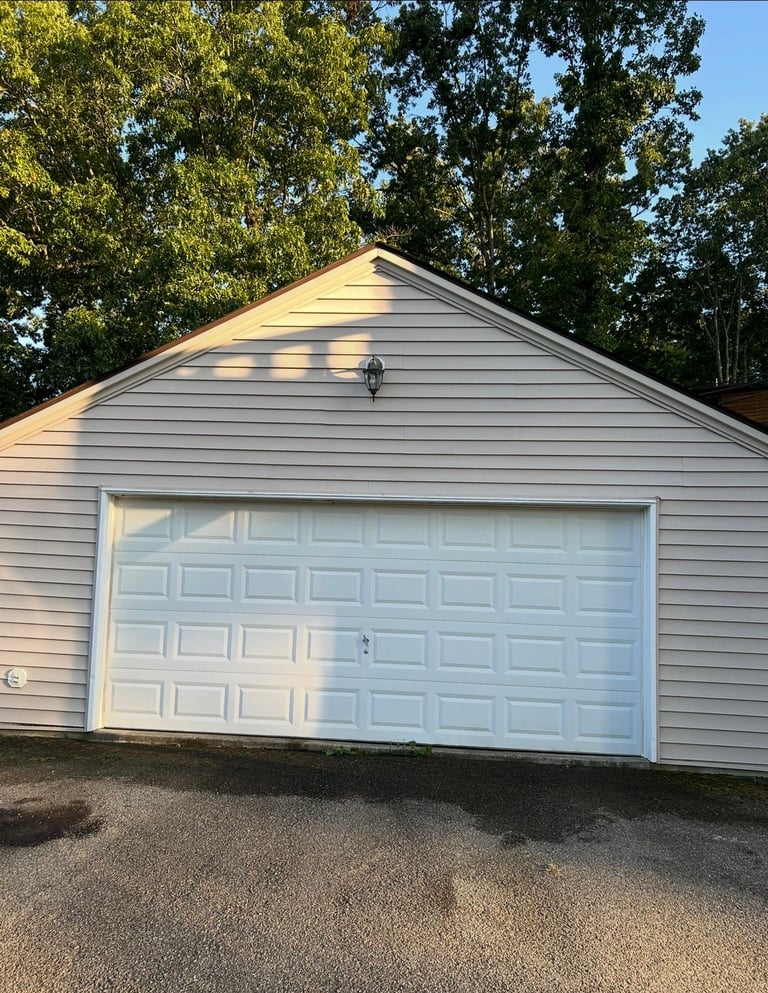 White garage with gabled roof surrounded by tall trees and a driveway