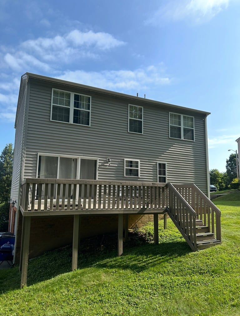 Elevated gray vinyl-sided house with wooden deck and stairs on a grassy lot under blue sky