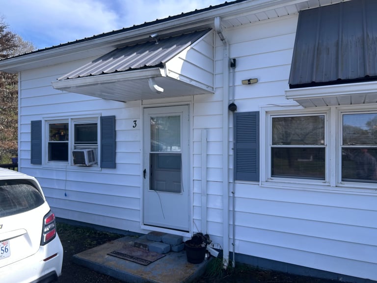 Light blue cottage with white trim dark shutters metal roof overhang and covered entrance with white car on left side