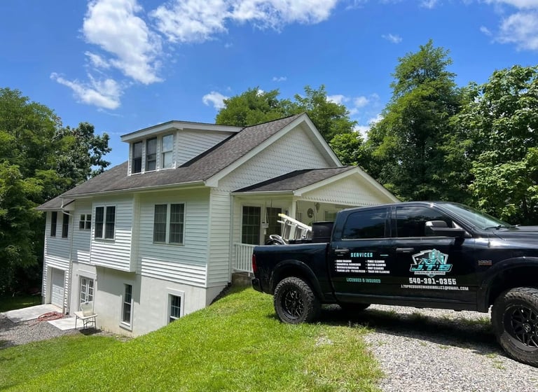 Elevated white two-story house with a black pickup truck parked in front on a gravel driveway, surrounded by green trees and lawn.