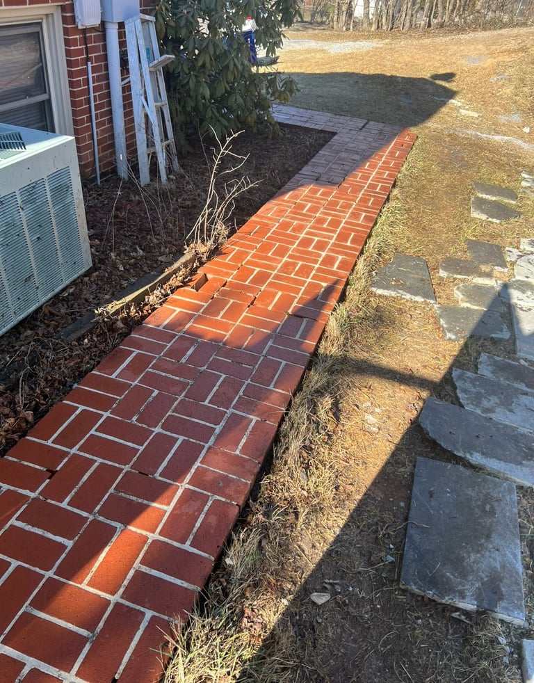 Red brick walkway along side of house with landscaping, shadows of trees and person visible on ground